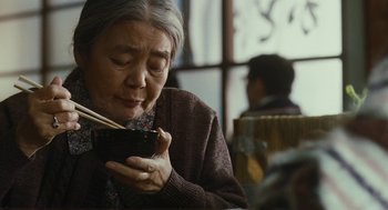 Movie still from “Shoplifters” (2018), directed by Kore-eda Hirokazu – An older woman holding chopsticks while eating a bowl of food; Close Up shot, Over the shoulder angle