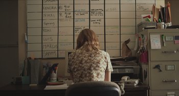 Movie still from “Short Term 12” (2013), directed by Destin Daniel Cretton – A woman sitting at a desk in front of a wall of post - it notes; Medium shot, Over the shoulder angle