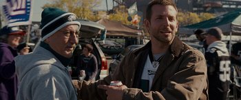 Movie still from “Silver Linings Playbook” (2012), directed by David O. Russell – A man holding a can of soda while standing next to another man; Medium shot, Over the shoulder angle