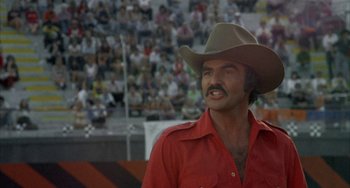 Movie still from “Smokey and the Bandit” (1977), directed by Hal Needham – A man in a red shirt and a cowboy hat is in front of an audience; Close Up shot, Low angle