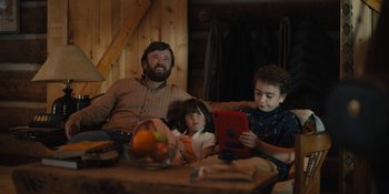 Movie still from “Somebody I Used to Know” (2023), directed by Dave Franco – A man and two children sitting on a couch looking at an ipad; Medium shot, Over the shoulder angle