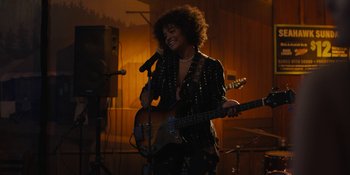 Movie still from “Somebody I Used to Know” (2023), directed by Dave Franco – A woman holding a microphone while holding a guitar; Medium shot, Over the shoulder angle