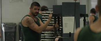 Movie still from “Son of a Gun” (2014), directed by Julius Avery – A man in a green tank top is holding a metal object; Medium shot, Over the shoulder angle
