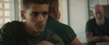 Movie still from “Son of a Gun” (2014), directed by Julius Avery – A young man with a beard and a green t - shirt is looking at the camera; Close Up shot, Over the shoulder angle