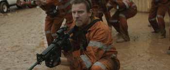 Movie still from “Son of a Gun” (2014), directed by Julius Avery – A man in an orange suit is holding a rifle; Medium shot, Over the shoulder angle