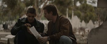 Movie still from “Son of a Gun” (2014), directed by Julius Avery – A man sitting next to another man holding papers; Medium shot, Over the shoulder angle