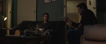 Movie still from “Son of a Gun” (2014), directed by Julius Avery – A man sitting on a couch holding a gun in his hand; Medium shot, Over the shoulder angle