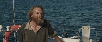 Movie still from “Son of a Gun” (2014), directed by Julius Avery – A man with long hair and a goatee is sitting on a sailboat; Medium shot, Low angle
