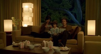 Movie still from “One Hour Photo” (2002), directed by Mark Romanek – Three people are sitting on a couch with a bowl on the table in front of them; Medium shot, High angle