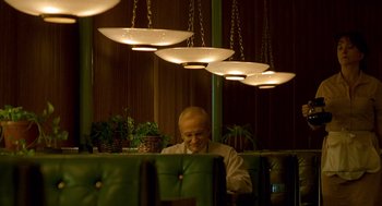 Movie still from “One Hour Photo” (2002), directed by Mark Romanek – A man sitting at a table in front of a bunch of lights; Medium shot, Low angle