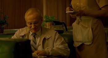 Movie still from “One Hour Photo” (2002), directed by Mark Romanek – An older man sitting at a table writing on a paper; Medium shot, Low angle