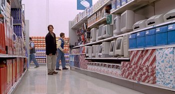 Movie still from “One Hour Photo” (2002), directed by Mark Romanek – Two people standing in front of a shelf of computers; Wide shot, Over the shoulder angle