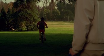 Movie still from “One Hour Photo” (2002), directed by Mark Romanek – A person riding a bike on a lush green field; Extreme Wide shot, Over the shoulder angle