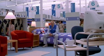 Movie still from “One Hour Photo” (2002), directed by Mark Romanek – A man sitting on top of a bed inside of a store; Wide shot, High angle