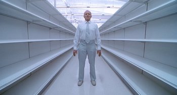Movie still from “One Hour Photo” (2002), directed by Mark Romanek – A man in a white suit and tie standing in an aisle of empty shelves; Wide shot, Low angle