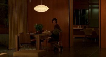 Movie still from “One Hour Photo” (2002), directed by Mark Romanek – A woman sitting at a table in front of a lamp; Wide shot, High angle