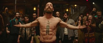 Movie still from “Sonic the Hedgehog 2” (2022), directed by Jeff Fowler – A man with a tattoo on his chest is standing in front of a group of men; Medium shot, Low angle