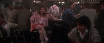 Movie still from “St. Elmo's Fire” (1985), directed by Joel Schumacher – A woman sitting on a bar in front of other people; Medium shot, Over the shoulder angle