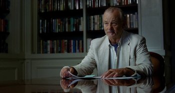 Movie still from “St. Vincent” (2014), directed by Theodore Melfi – An older man sitting at a table in front of a bookshelf; Medium shot, Over the shoulder angle