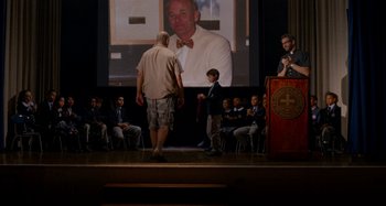 Movie still from “St. Vincent” (2014), directed by Theodore Melfi – A man standing on a stage next to a man in front of an audience; Wide shot, Over the shoulder angle