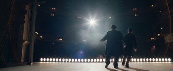 Movie still from “Stan & Ollie” (2018), directed by Jon S. Baird – A man wearing a hat and a black coat walking across a stage; Wide shot, Low angle