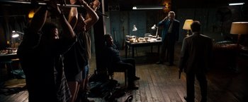 Movie still from “Stand Up Guys” (2012), directed by Fisher Stevens – A group of people standing in a room with a man sitting in a chair; Wide shot, High angle