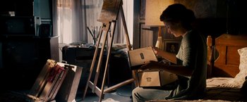 Movie still from “Stand Up Guys” (2012), directed by Fisher Stevens – A person sitting in front of an easel holding boxes; Medium shot, High angle
