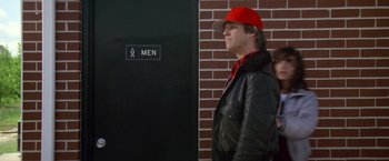 Movie still from “Starman” (1984), directed by John Carpenter – A man wearing a red hat standing in front of a men's restroom; Medium shot, Low angle