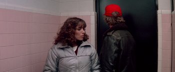 Movie still from “Starman” (1984), directed by John Carpenter – A woman in a silver jacket standing next to a man in a red hat; Medium shot, Over the shoulder angle