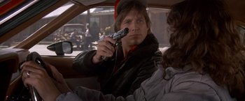 Movie still from “Starman” (1984), directed by John Carpenter – A man holding a gun in a car while sitting in the passenger seat; Close Up shot, Over the shoulder angle