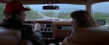 Movie still from “Starman” (1984), directed by John Carpenter – Two people sitting in the drivers seat of a car on the road; Medium shot, High angle