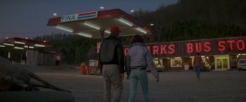 Movie still from “Starman” (1984), directed by John Carpenter – A man and a woman holding hands in front of a gas station at night; Wide shot, Over the shoulder angle