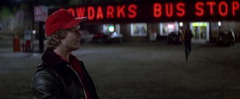 Movie still from “Starman” (1984), directed by John Carpenter – A man wearing a red hat standing in front of a building; Medium shot, Over the shoulder angle