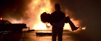 Movie still from “Starman” (1984), directed by John Carpenter – A man holding a woman in front of a burning building; Wide shot, Low angle