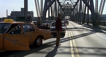 Movie still from “Stripes” (1981), directed by Ivan Reitman – A man standing on the side of a bridge next to a line of parked cars; Wide shot, Over the shoulder angle
