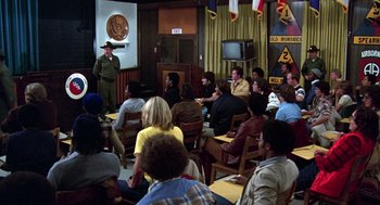 Movie still from “Stripes” (1981), directed by Ivan Reitman – A group of people sitting in front of a man in a hat; Wide shot, High angle
