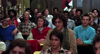 Movie still from “Stripes” (1981), directed by Ivan Reitman – A group of people sitting in front of a microphone; Medium shot, Over the shoulder angle