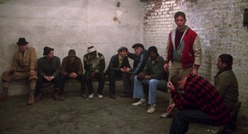 Movie still from “Stripes” (1981), directed by Ivan Reitman – A group of men sitting on a bench in front of a brick wall; Wide shot, High angle