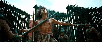 Movie still from “Ong Bak 3” (2010), directed by Tony Jaa – A man holding a wooden stick in the middle of a scene; Medium shot, Low angle