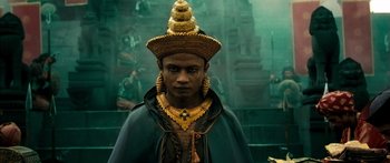 Movie still from “Ong Bak 3” (2010), directed by Tony Jaa – A man wearing a gold hat and a green cape; Close Up shot, High angle
