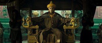 Movie still from “Ong Bak 3” (2010), directed by Tony Jaa – A man sitting on top of a chair wearing a hat; Wide shot, Low angle