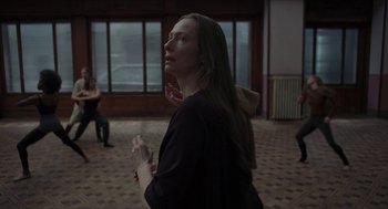 Movie still from “Suspiria” (2018), directed by Luca Guadagnino – A woman standing in front of a window with her hands clasped together; Medium shot, Over the shoulder angle