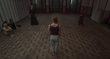 Movie still from “Suspiria” (2018), directed by Luca Guadagnino – A woman standing in front of two other women in an empty room; Wide shot, High angle
