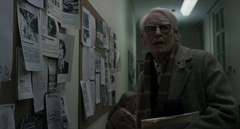 Movie still from “Suspiria” (2018), directed by Luca Guadagnino – An old man wearing glasses and a scarf is standing in front of a bulletin board; Medium shot, Over the shoulder angle