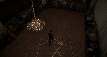 Movie still from “Suspiria” (2018), directed by Luca Guadagnino – A man standing in a room under a chandelier; Extreme Wide shot, Overhead angle