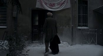Movie still from “Suspiria” (2018), directed by Luca Guadagnino – A man walking in front of a building with a sign on it; Wide shot, Over the shoulder angle