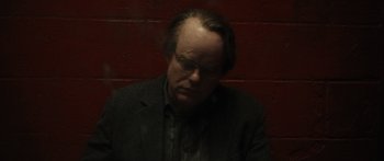 Movie still from “Synecdoche, New York” (2008), directed by Charlie Kaufman – A man in a suit and glasses is sitting in front of a brick wall; Close Up shot, High angle