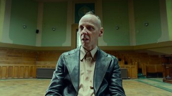 Movie still from “T2 Trainspotting” (2017), directed by Danny Boyle – A bald man in a leather jacket sitting in front of a church; Close Up shot, Low angle