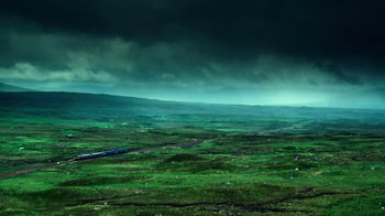 Movie still from “T2 Trainspotting” (2017), directed by Danny Boyle – A train traveling through a lush green valley under a cloudy sky; Extreme Wide shot, High angle