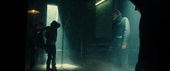 Movie still from “Taken 2” (2012), directed by Olivier Megaton – A man standing next to another man in a dark room; Wide shot, Over the shoulder angle